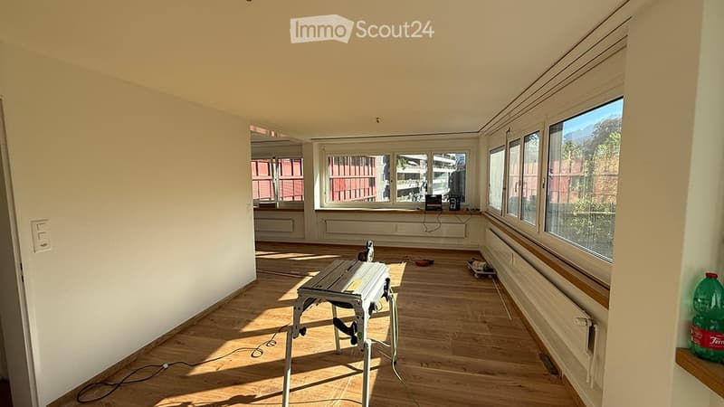 Empty room with wooden flooring, large windows, and a workbench in the center. No furniture or personal items are present.