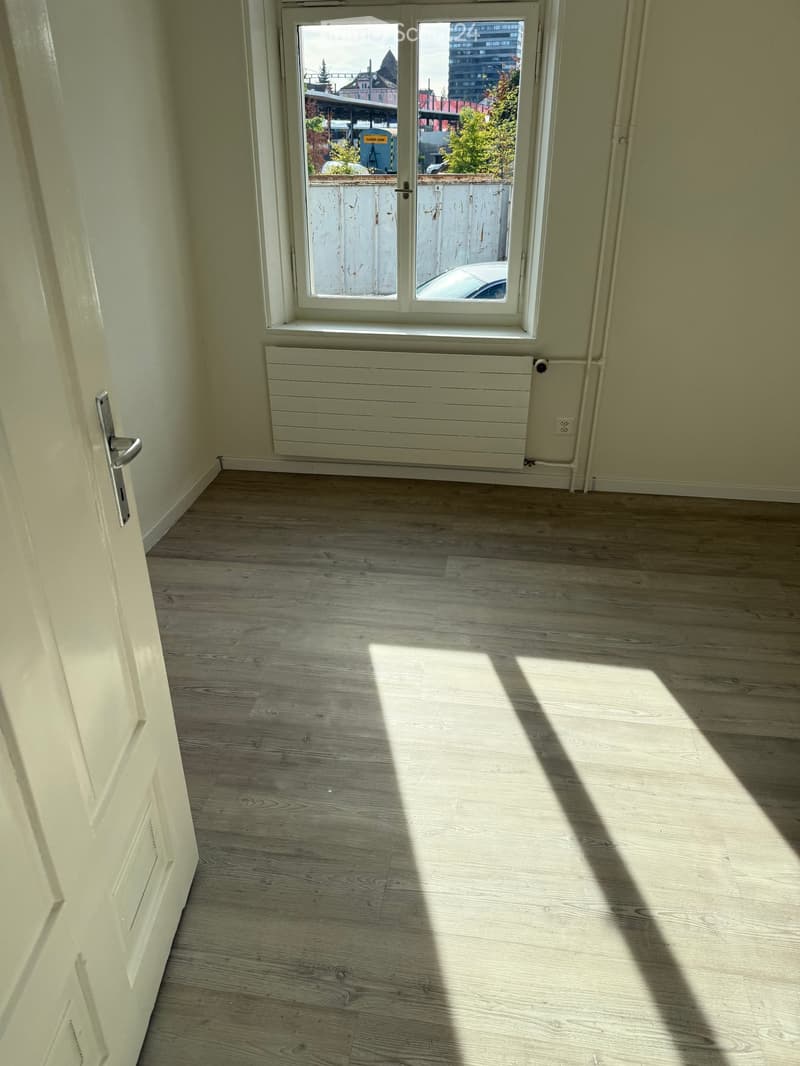 Empty room with light wooden flooring, white walls, a door on the left, and a window with a radiator below it. Sunlight streams in, casting shadows on the floor.