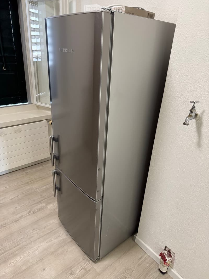 Modern stainless steel refrigerator in a kitchen with light wooden flooring and white walls.