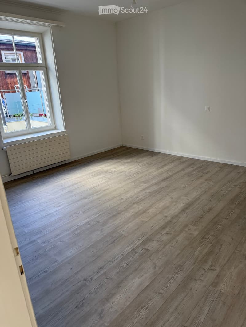 Empty room with light wooden flooring, white walls, and a large window with a radiator below it.