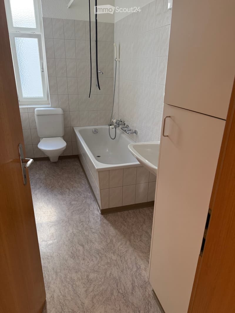 Bathroom with a bathtub, toilet, sink, and shower hose. Light-colored tiles and a carpeted floor.