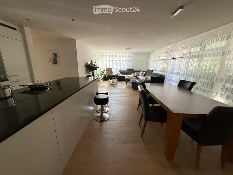 Open-plan living and dining area with modern furniture, including a sofa, coffee table, dining table with chairs, and a kitchen counter with an induction stove and frying pan. The space features wooden flooring and large windows with sheer curtains.