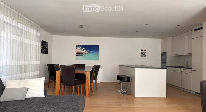 Open-plan living and dining area with a grey sofa, dining table with six chairs, kitchen island with stools, and modern cabinets.