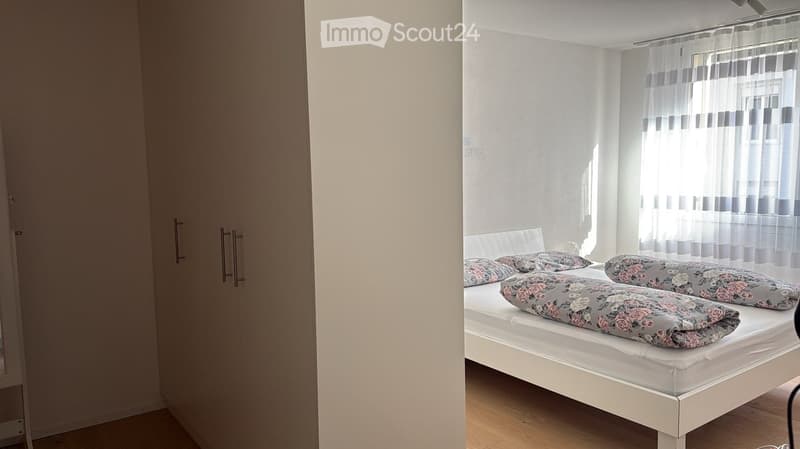 A tidy bedroom with a white bed, floral pillows, and a large wardrobe. Natural light filters through sheer curtains.