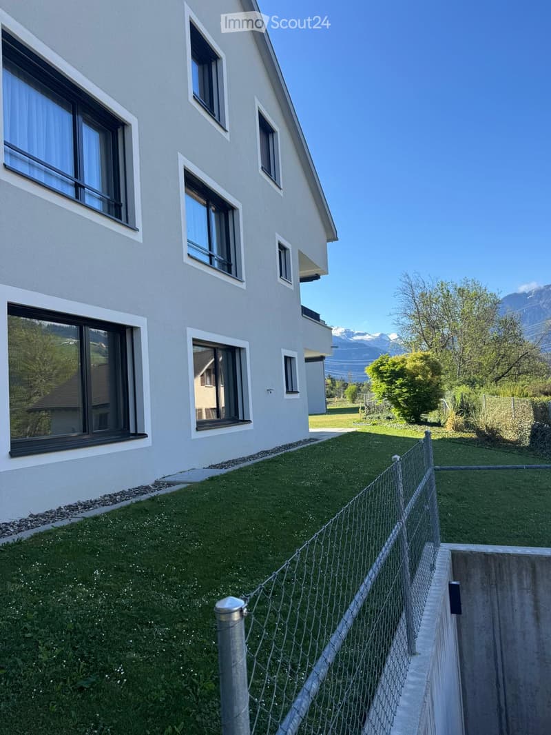Modern two-story house with light gray exterior, multiple large windows, and a well-maintained lawn enclosed by a wire fence. The background features a scenic mountain view.
