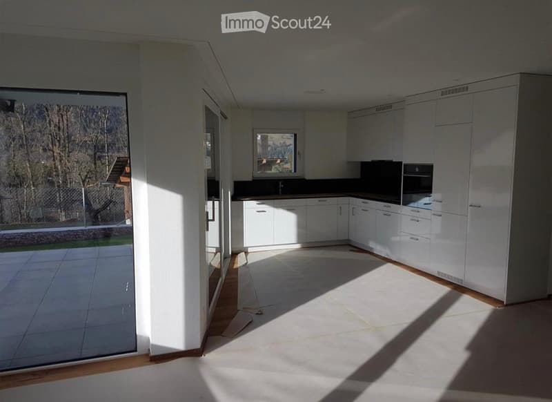 Empty kitchen with white cabinets, a countertop, and a large window letting in natural light. The floor is tiled, and there is a glass door leading to an outdoor area.