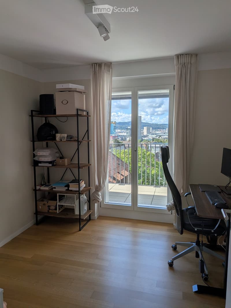 Bright room with wooden floor, large window with view, desk, chair, shelf with books and items.