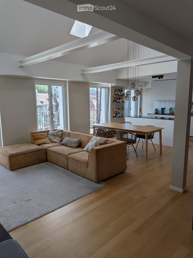 Open-plan living and dining area with wooden flooring, a sectional sofa, a dining table with chairs, and a modern kitchen area with pendant lights and shelves.