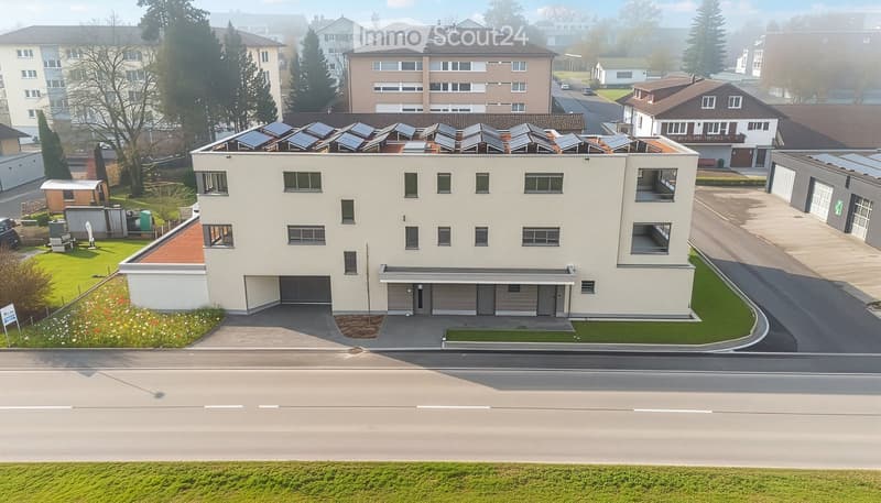 White multi-story building with solar panels on the roof, 3 garages on the ground floor, multiple windows, paved driveway, gardens and grass around, several houses in the background.