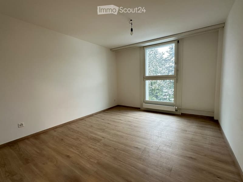 Empty room with wooden flooring, white walls, and a large window with a view of trees.