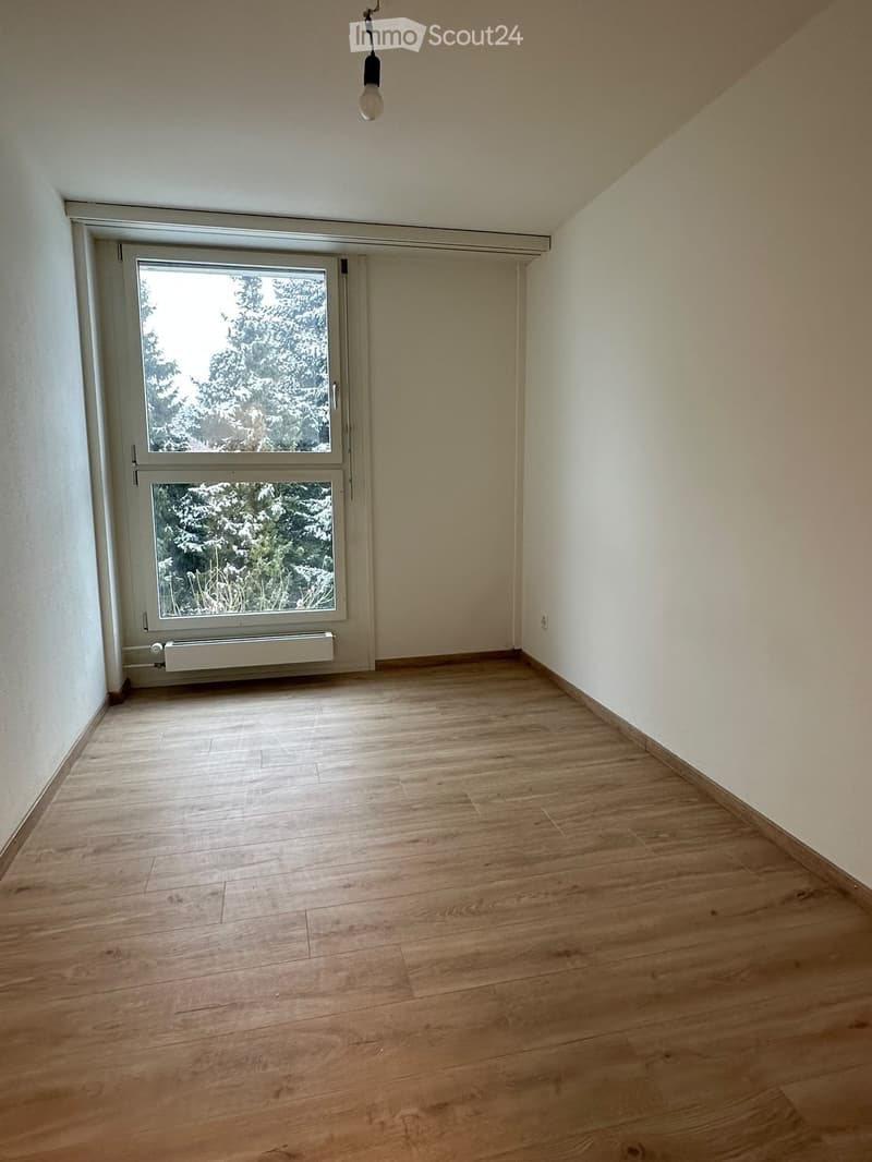 Empty room with wooden flooring, white walls, and a large window with a view of trees.