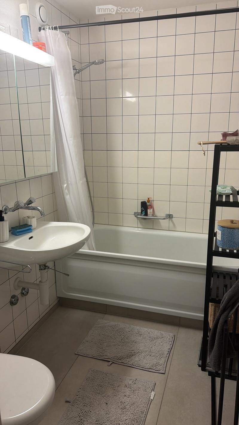 Bathroom with white tiled walls, a bathtub with a shower curtain, a sink with a faucet, a toilet, and a small shelf holding toiletries. Two grey rugs are placed on the floor near the bathtub.