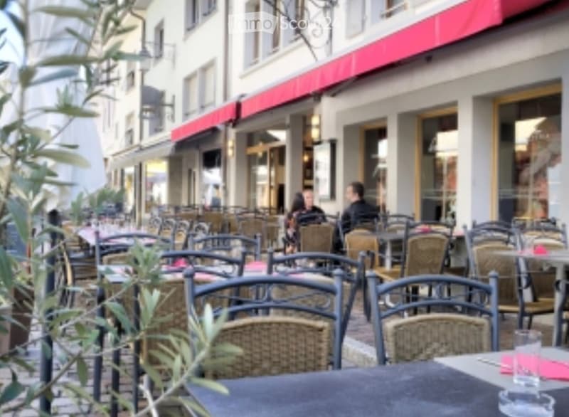 An outdoor seating area with tables and chairs in front of a building with a red awning. The area is surrounded by greenery and has a paved surface.