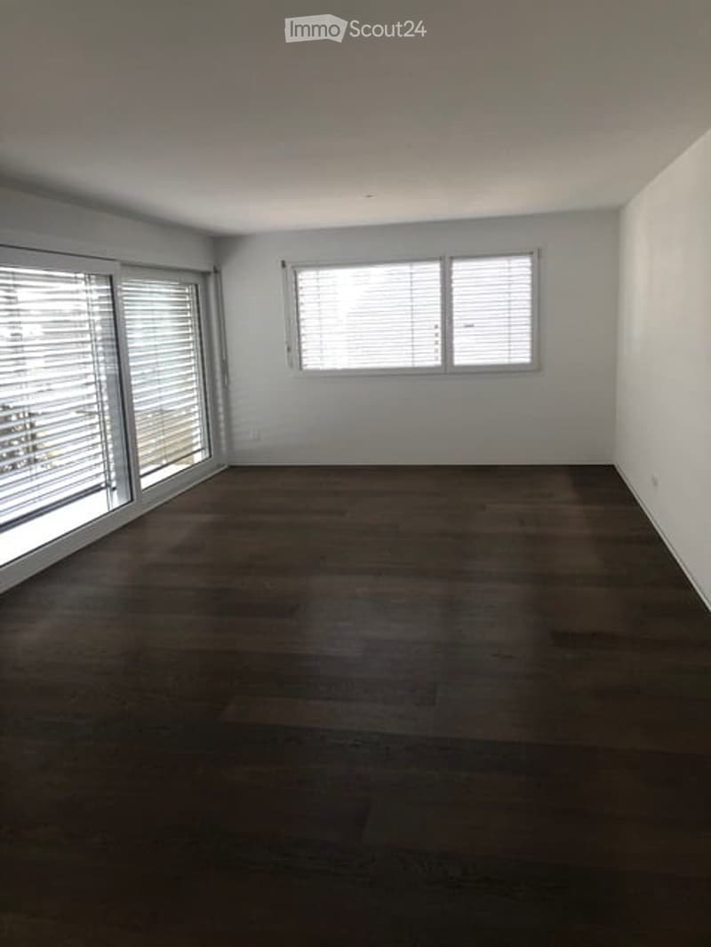 Completely empty room with wooden flooring, white walls, and large windows with blinds.