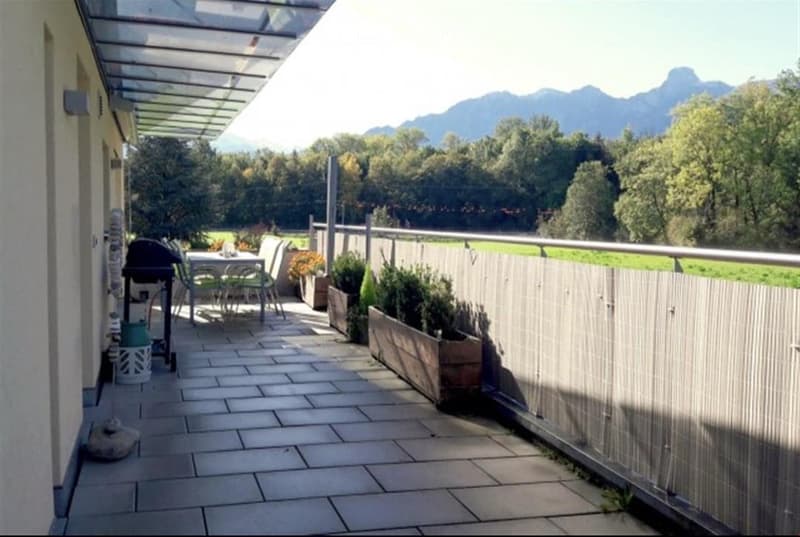 Balcony with tiled floor, metal railing, outdoor furniture including a table, chairs, and a barbecue, with potted plants and a scenic mountain view.