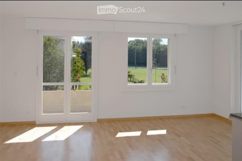 Empty room with white walls, wooden floor, large windows, and sliding glass doors leading to an outdoor area.