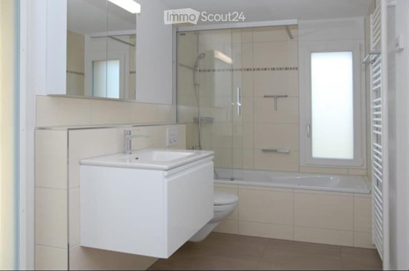 Modern bathroom with a white sink, mirror, toilet, bathtub, and glass-enclosed shower. Light-colored tiles and a radiator are visible.