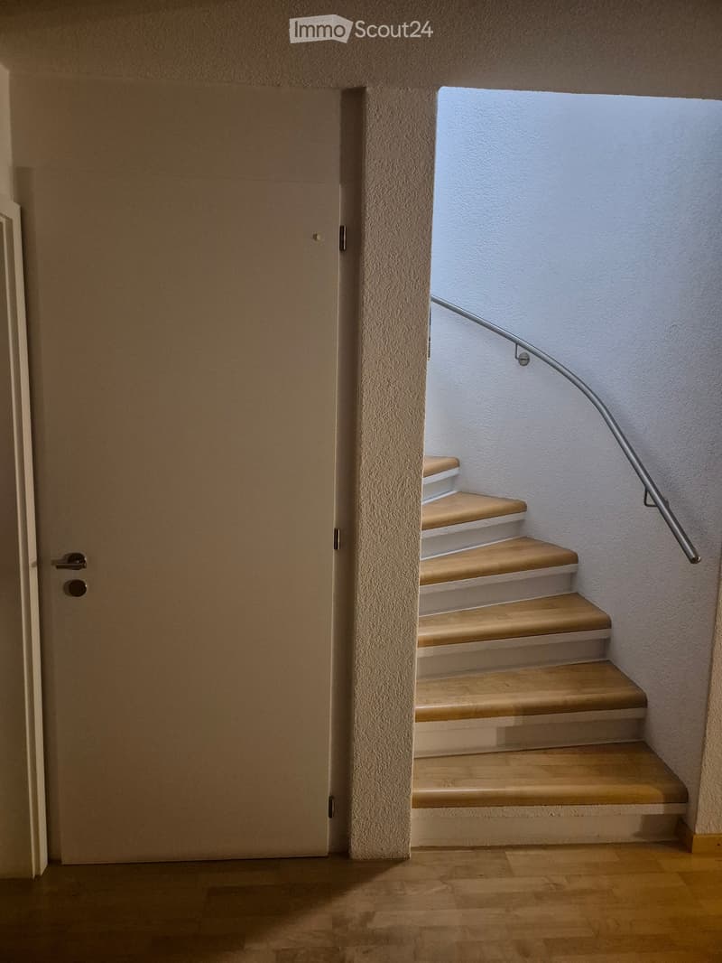 A modern hallway with a wooden floor, a closed door on the left, and a staircase with wooden steps and a metal handrail leading upwards.