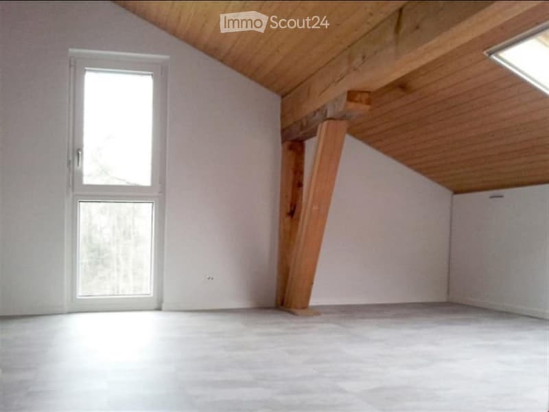 Empty attic space with wooden beams, white walls, and a large window allowing natural light. The floor is covered with a smooth, light-colored material.