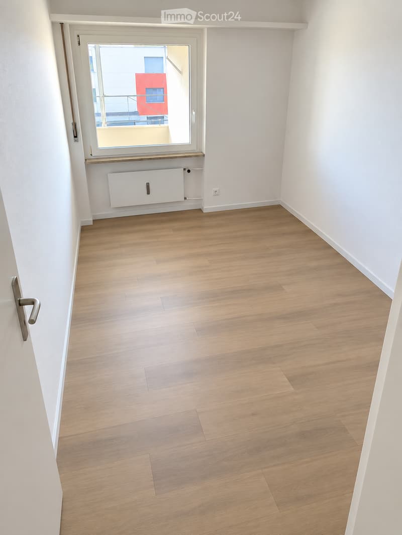 Empty room with light wooden flooring, white walls, and a large window with a view of a red building and balcony area.