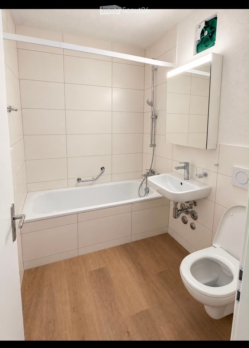 Modern bathroom with a bathtub, sink, toilet, and mirror. Features light-colored tiles and wooden flooring.