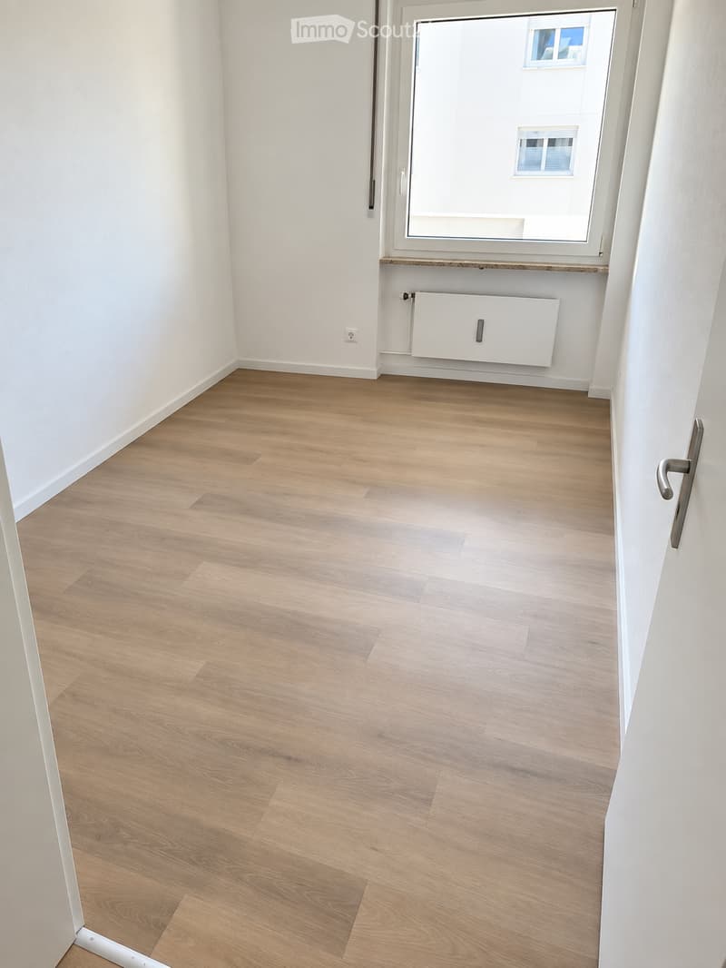 Empty room with light wooden flooring, white walls, a window with a radiator below, and a door handle visible on the right side.