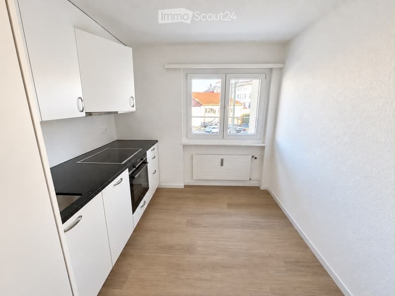 Modern kitchen with white cabinets, black countertop, and a built-in stove and oven. The room features light wooden flooring and large windows offering a view of the street.