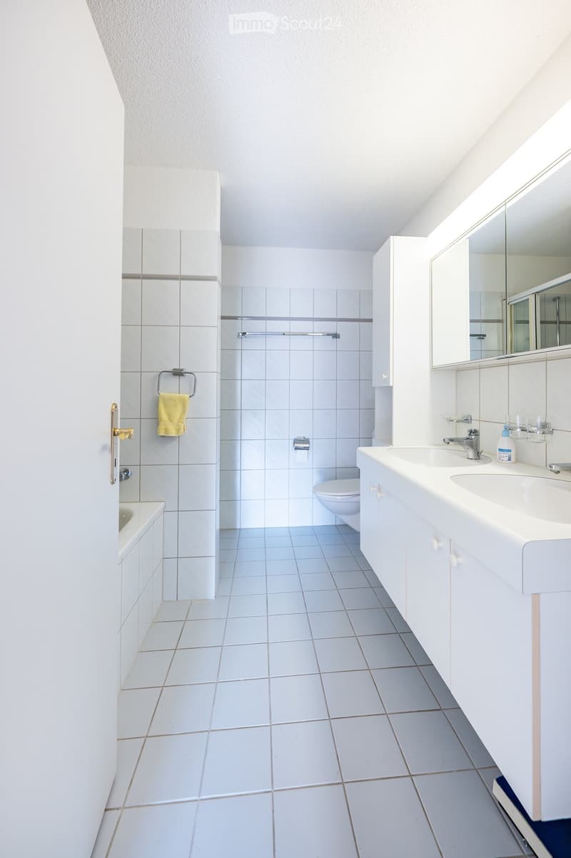 Modern bathroom with white tiles, double sink, mirror, toilet, and bathtub. Features a towel on a rack and a sleek design.