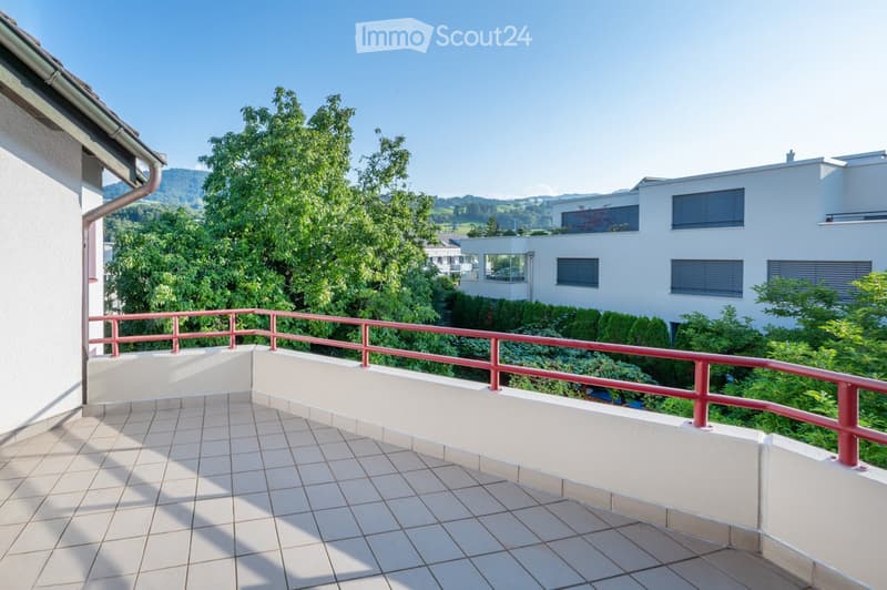 Balcony with tiled floor, red metal railing, overlooking greenery and distant mountains.