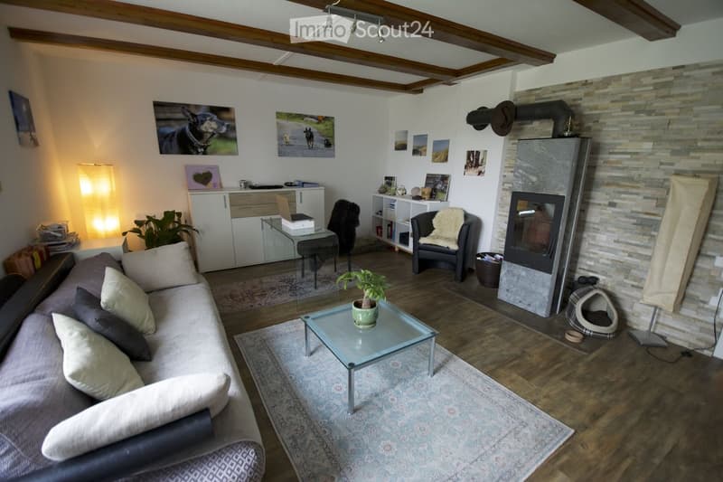Cozy living room with exposed wooden beams, a fireplace, a grey sofa with multiple cushions, a glass coffee table, an armchair with a fur throw, a laptop on a glass table, and decorative picture frames on the wall.
