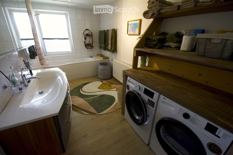 Bathroom with a sink, bathtub, washing machine, shelves with towels, a picture frame, and a basket on the floor.