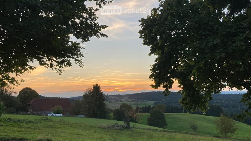 A serene rural landscape at sunset, featuring rolling green hills, scattered trees, and a small traditional house with a red-tiled roof in the distance.