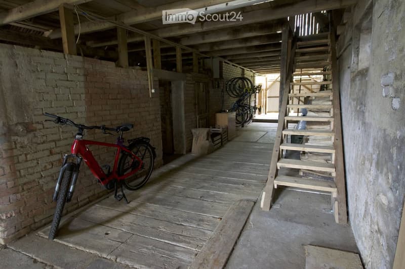 An unfinished wooden corridor with exposed brick walls, a wooden staircase leading upwards, and a red bicycle leaning against the wall.