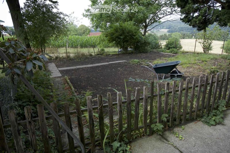 Garden area enclosed by a wooden fence, featuring a wheelbarrow, garden bed, and surrounding greenery with trees and open fields in the background.