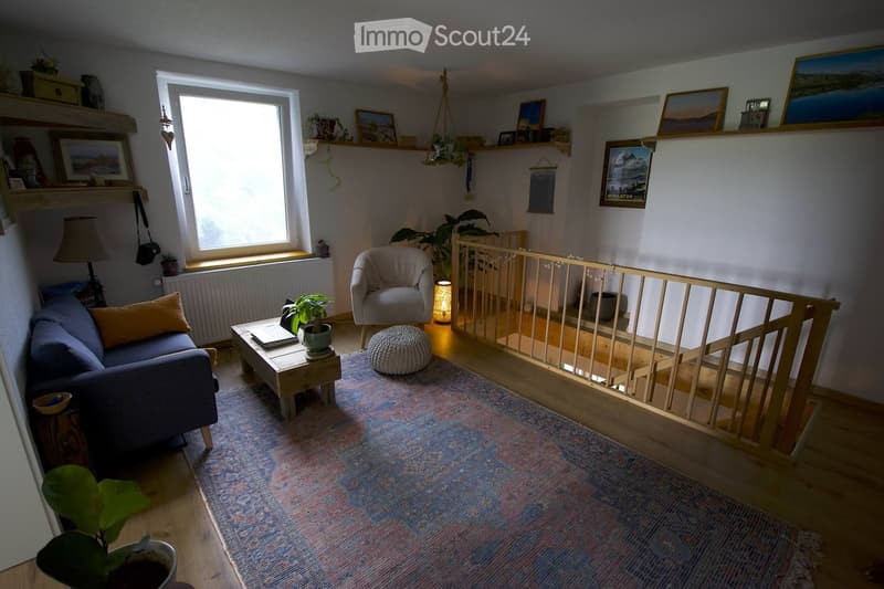 Cozy living room with a blue sofa, wooden coffee table, armchair, ottoman, and a colorful rug. Shelves hold picture frames and decorative items, with plants adding a touch of greenery.
