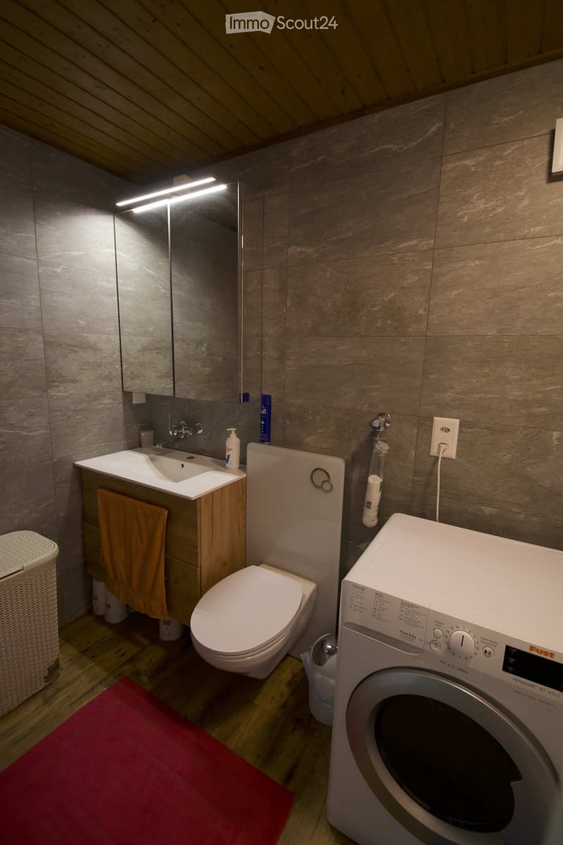 Modern bathroom with wooden floor, grey tiled walls, sink, toilet, washing machine, and a wicker basket. A towel hangs on the cabinet, and a mirror is mounted on the wall.
