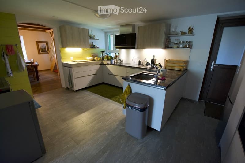 Modern kitchen with light-colored cabinets, a central island, and a sink with a faucet. The countertop is cluttered with various items, including a trash bin, a microwave, and a refrigerator. The floor is tiled, and the walls are painted in a light color.