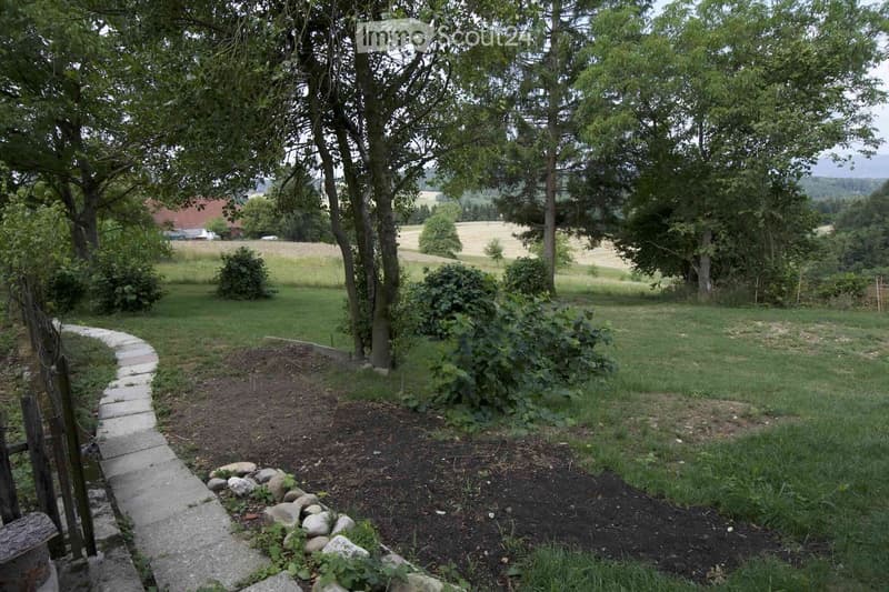 Garden area with a stone pathway, trees, bushes, and a view of a field and distant house.