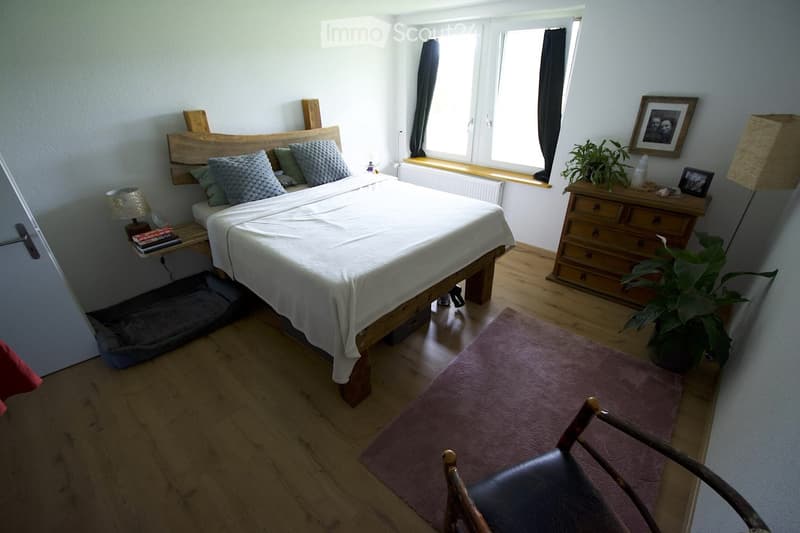 Cozy bedroom with wooden flooring, a large bed with a wooden headboard, nightstand with a lamp, a wooden chair, a potted plant, a dresser with drawers, and a cat bed.