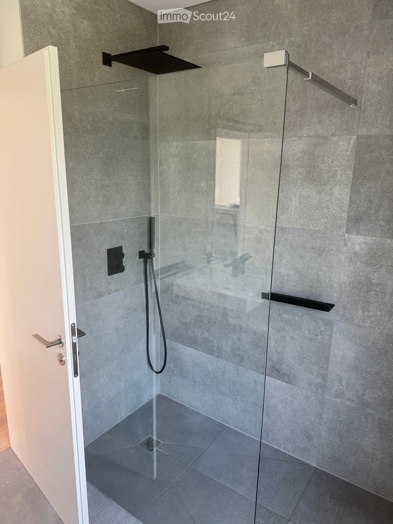 Modern shower area with a glass partition, grey tiled walls, and a sleek black shower handle.