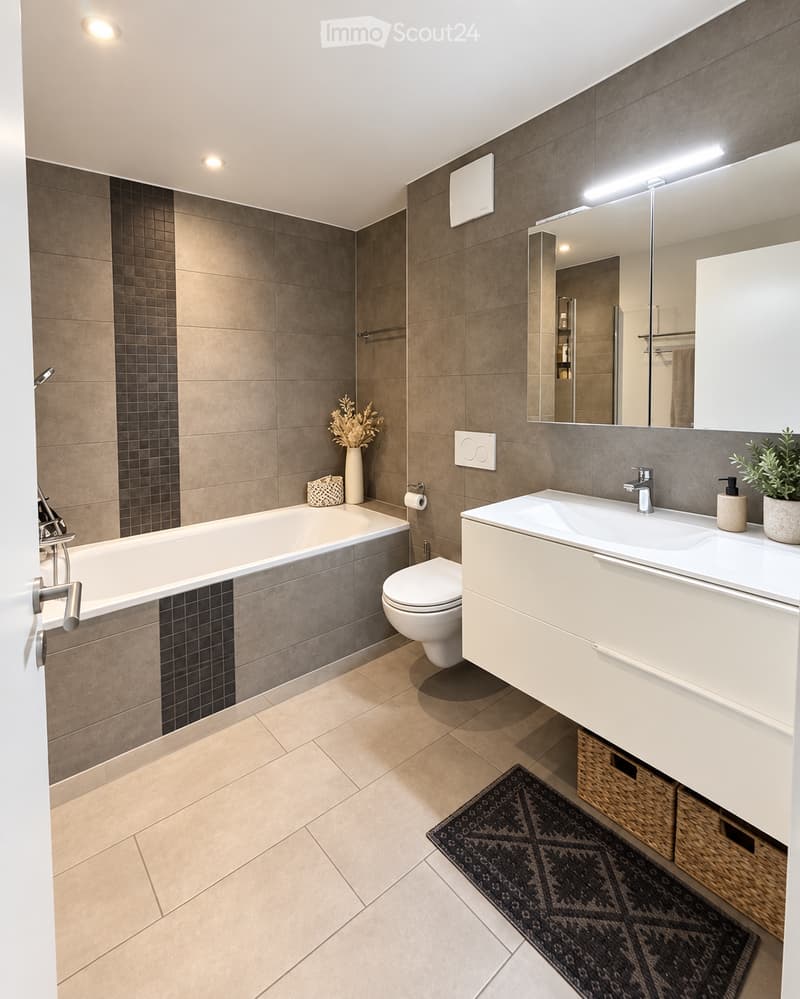 Modern bathroom with a bathtub, toilet, sink, large mirror, and decorative vase on the edge of the tub. The space features beige tiles, a wicker basket under the sink, and a patterned rug on the floor.