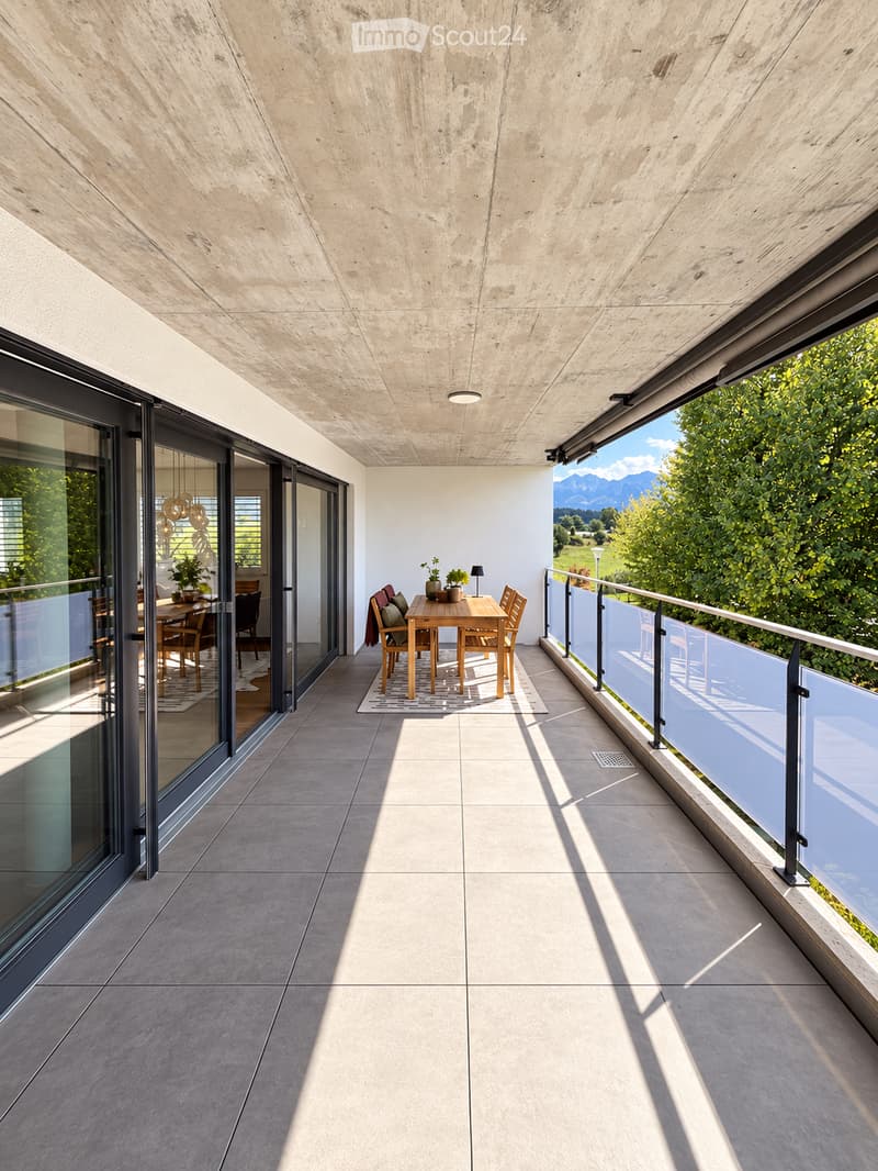 Modern balcony with concrete ceiling, tiled floor, glass railing, and a small dining set with a table, chairs, and potted plants.