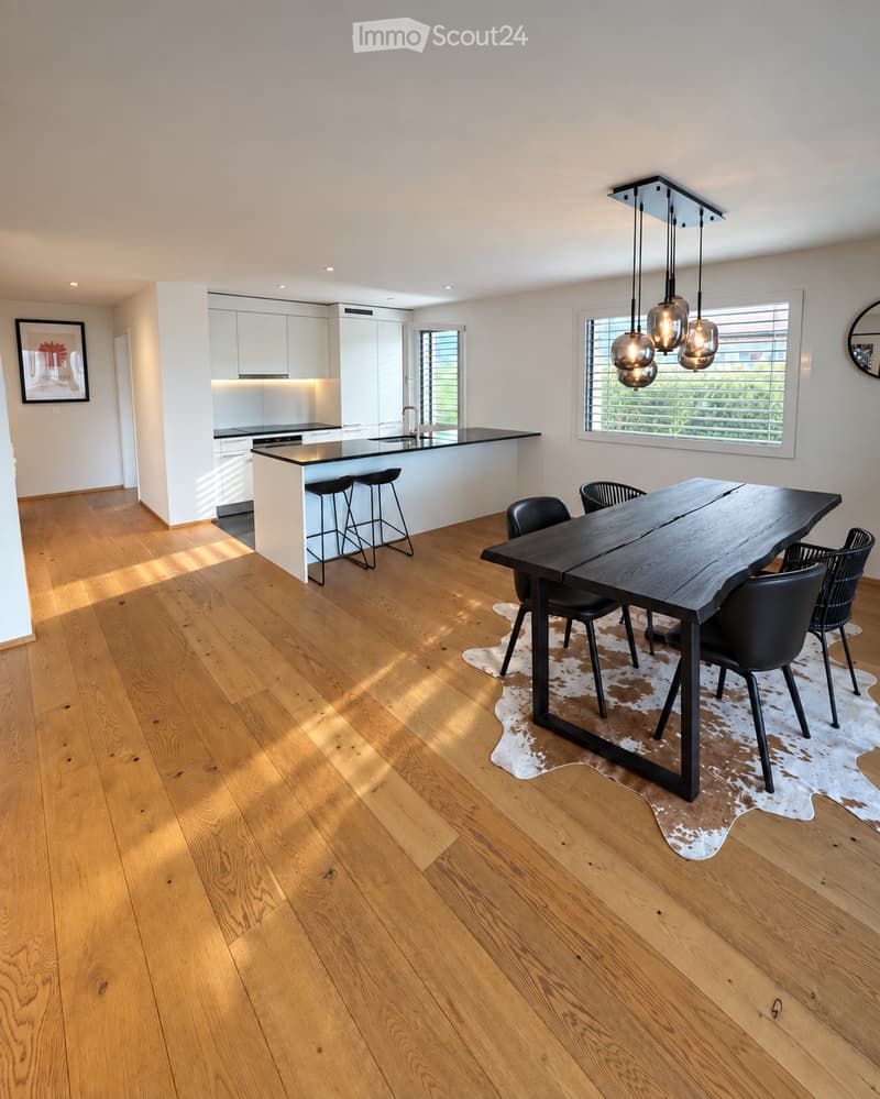 Open-plan kitchen and dining area with light wood flooring, white cabinetry, a black countertop, a large wooden dining table, and modern pendant lights.