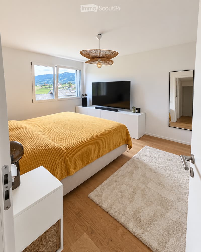 Modern bedroom with a large bed covered in a mustard-yellow duvet, a flat-screen TV on a white cabinet, a nightstand with a lamp, and a fluffy white rug on the wooden floor. Large windows offer a scenic mountain view.