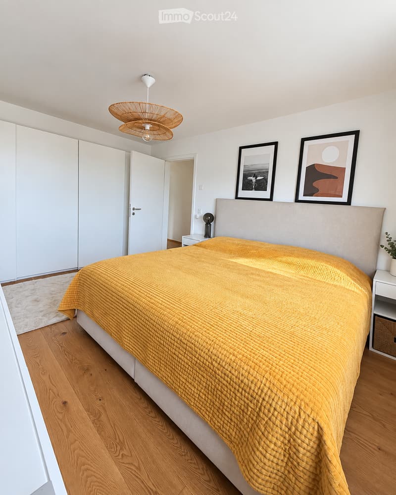 Modern bedroom with a large bed covered in a mustard yellow duvet, wooden flooring, white built-in wardrobes, and a wicker pendant light fixture.