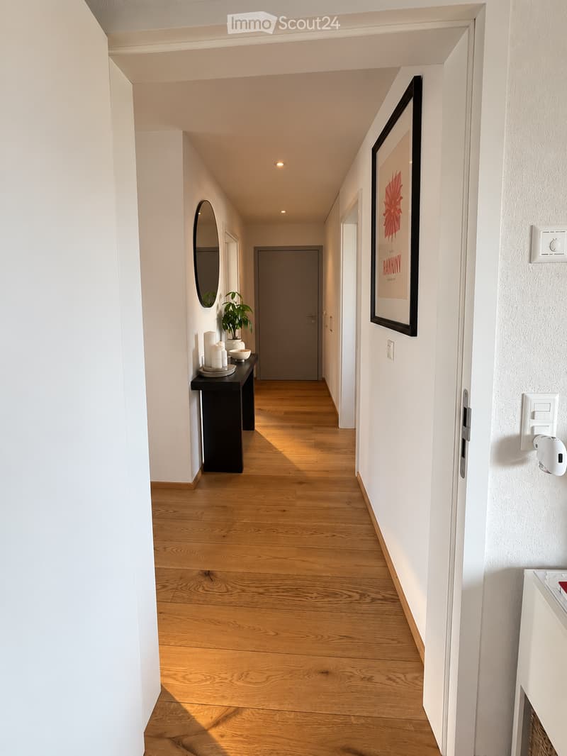 A modern hallway with light wooden flooring, white walls, and recessed lighting. Features a small black table with a plant and candles, a round mirror, and a framed picture on the wall.