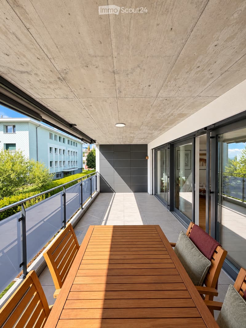 Modern balcony with a wooden table and chairs, glass railing, and a view of greenery and a building.