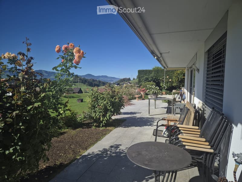 Balcony with a round table and several chairs, surrounded by greenery and a scenic mountain view.