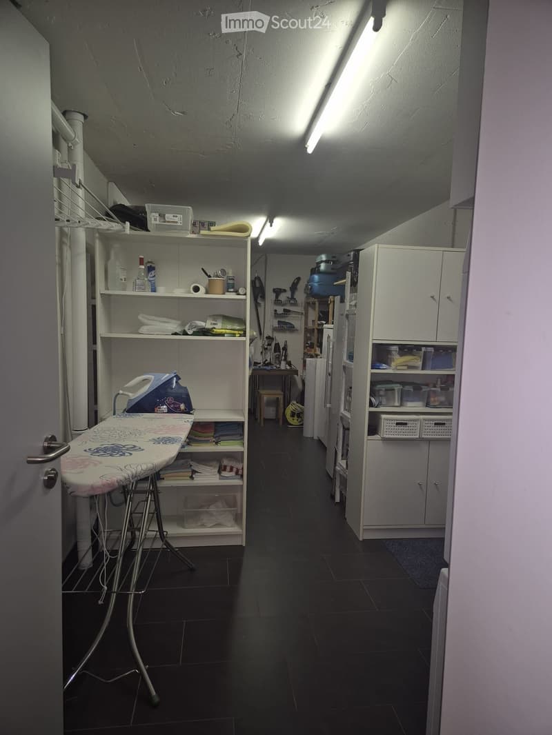 Laundry room with dark tiled floor, white walls, shelves filled with cleaning supplies, an ironing board, and a small table with cleaning tools.