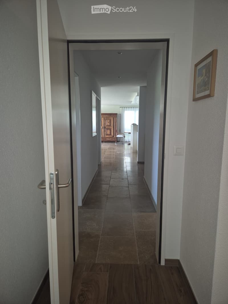 A long, narrow hallway with tiled flooring, white walls, and a wooden door at the end. A framed picture hangs on the right wall, and a partially visible room with furniture is seen through an open doorway.